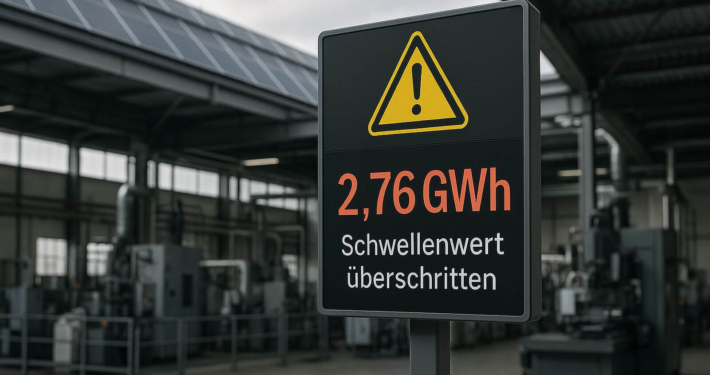 Warnschild mit der Anzeige 2,76 GWh und dem Hinweis „Schwellenwert überschritten“ vor einer Industriehalle mit Maschinen und Solaranlage