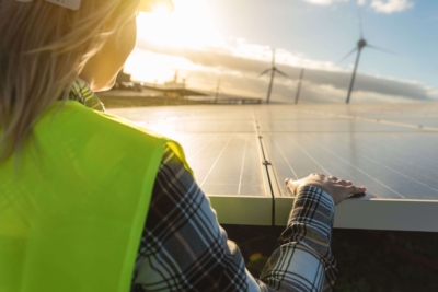 Energieberaterin mit Sicherheitsweste prüft Solaranlage bei Sonnenuntergang, Windkraftanlagen im Hintergrund