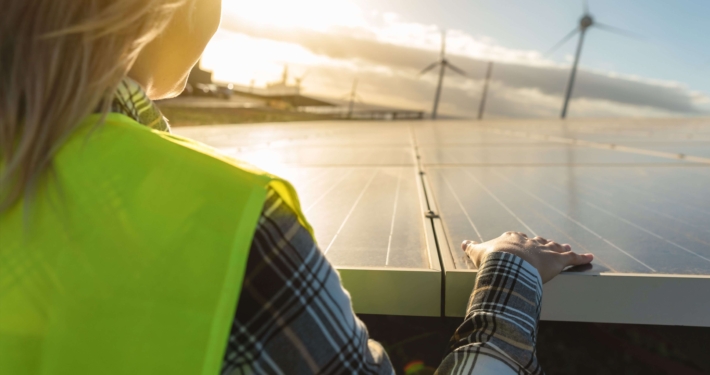 Energieberaterin mit Sicherheitsweste prüft Solaranlage bei Sonnenuntergang, Windkraftanlagen im Hintergrund