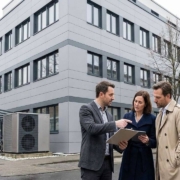 Ein professioneller Energieberater in Business-Kleidung steht vor einem modernen, grau fassadengedämmten Bürogebäude in Deutschland und erklärt zwei Entscheidern (Mann und Frau) anhand eines Tablets und Klemmbretts Sanierungspläne. Neben dem Gebäudeeingang ist eine große industrielle Wärmepumpe sichtbar installiert.