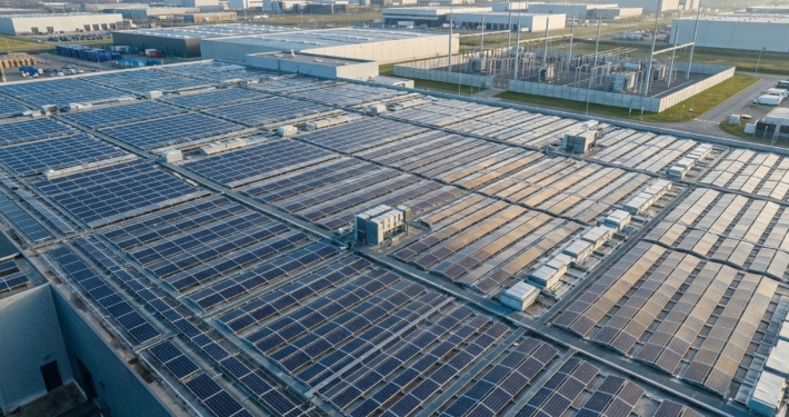 Großflächige Photovoltaik-Anlage auf einem Industriedach zur Eigenstromversorgung, im Hintergrund Windräder und ein Umspannwerk.