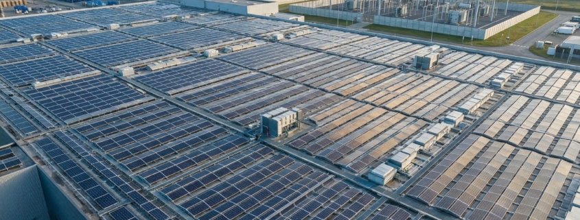 Großflächige Photovoltaik-Anlage auf einem Industriedach zur Eigenstromversorgung, im Hintergrund Windräder und ein Umspannwerk.
