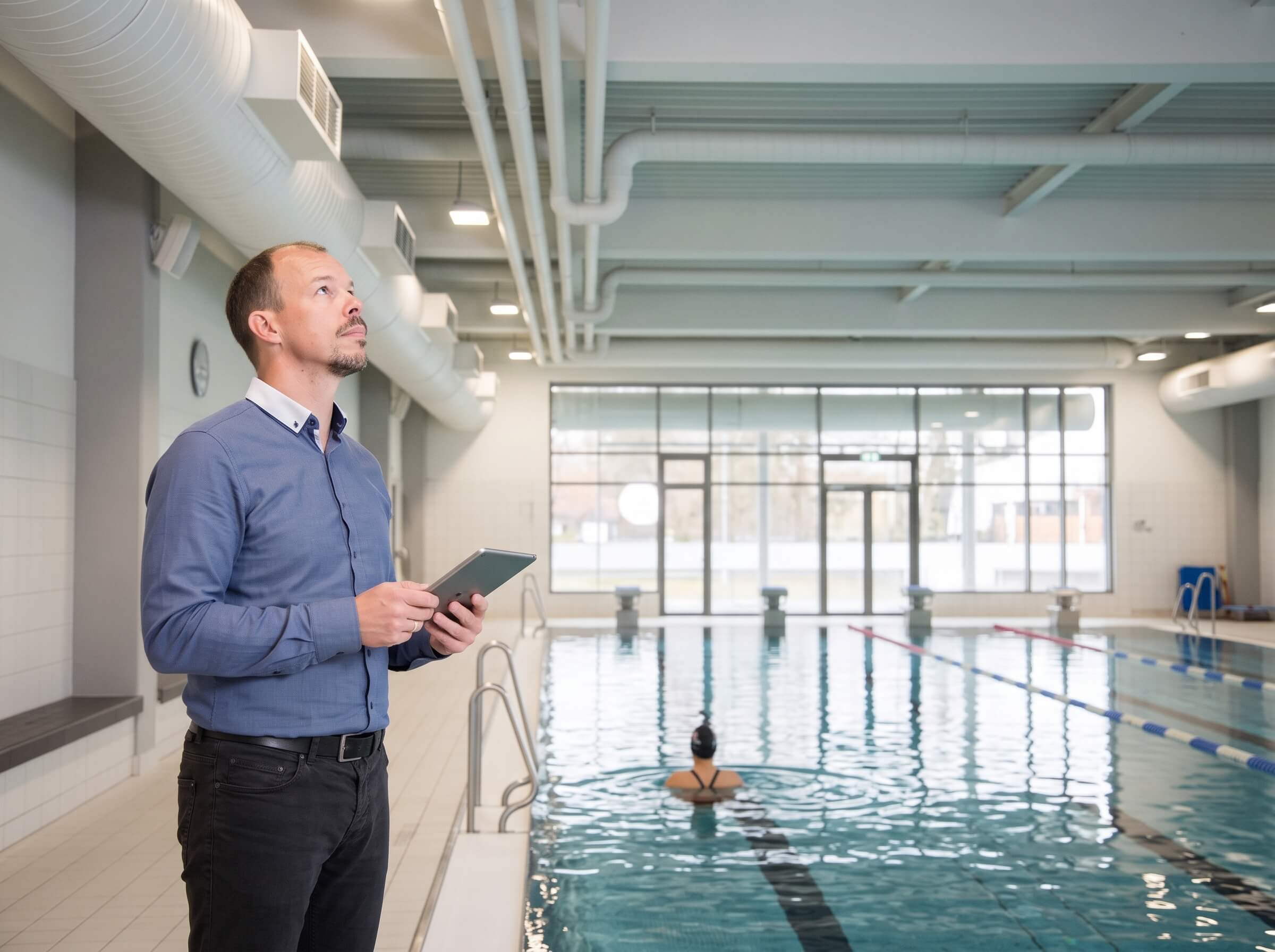 Fachplaner mit Tablet am Beckenrand einer Schwimmhalle, Blick zur Lüftungstechnik unter der Decke