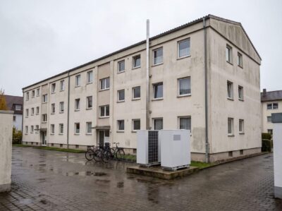 Wohnanlage aus den 1970er-Jahren mit Abgasrohr auf dem Dach und zwei Wärmepumpen-Außeneinheiten im Innenhof bei Nieselregen