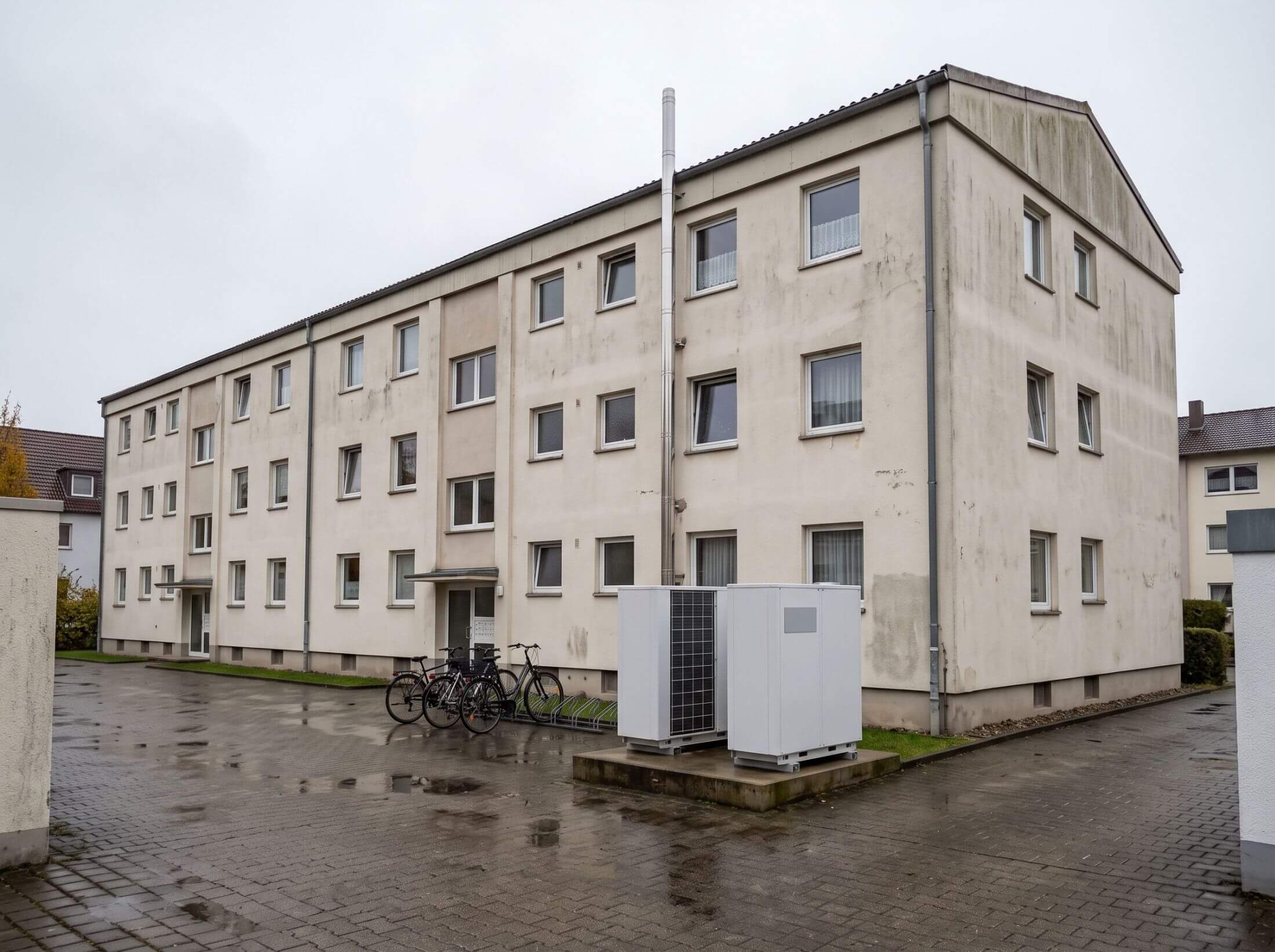 Wohnanlage aus den 1970er-Jahren mit Abgasrohr auf dem Dach und zwei Wärmepumpen-Außeneinheiten im Innenhof bei Nieselregen